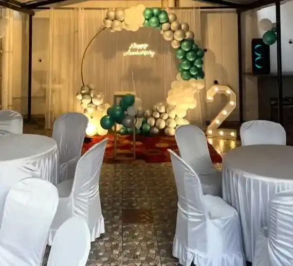 A green, cream and gold balloon arch around a Happy Anniversary neon sign, a lit number two standing beside it and banquet tables in white covers