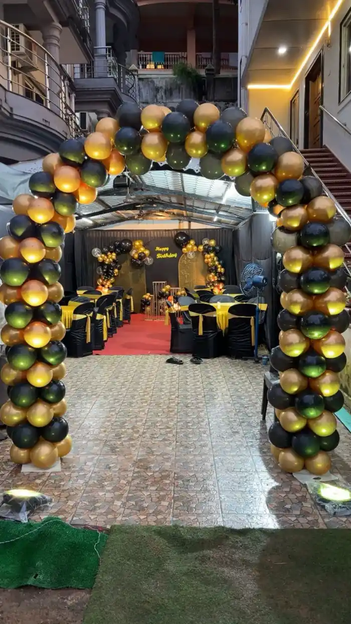 A black and gold balloon archway framing the entrance to a covered party space, with a Happy Birthday backdrop and dressed tables at the far end
