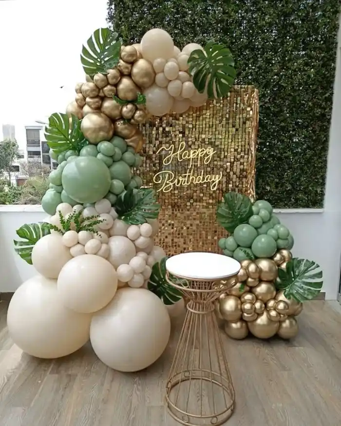 A balloon garland in sage, cream and gold with monstera leaves, framing a gold sequin wall reading Happy Birthday