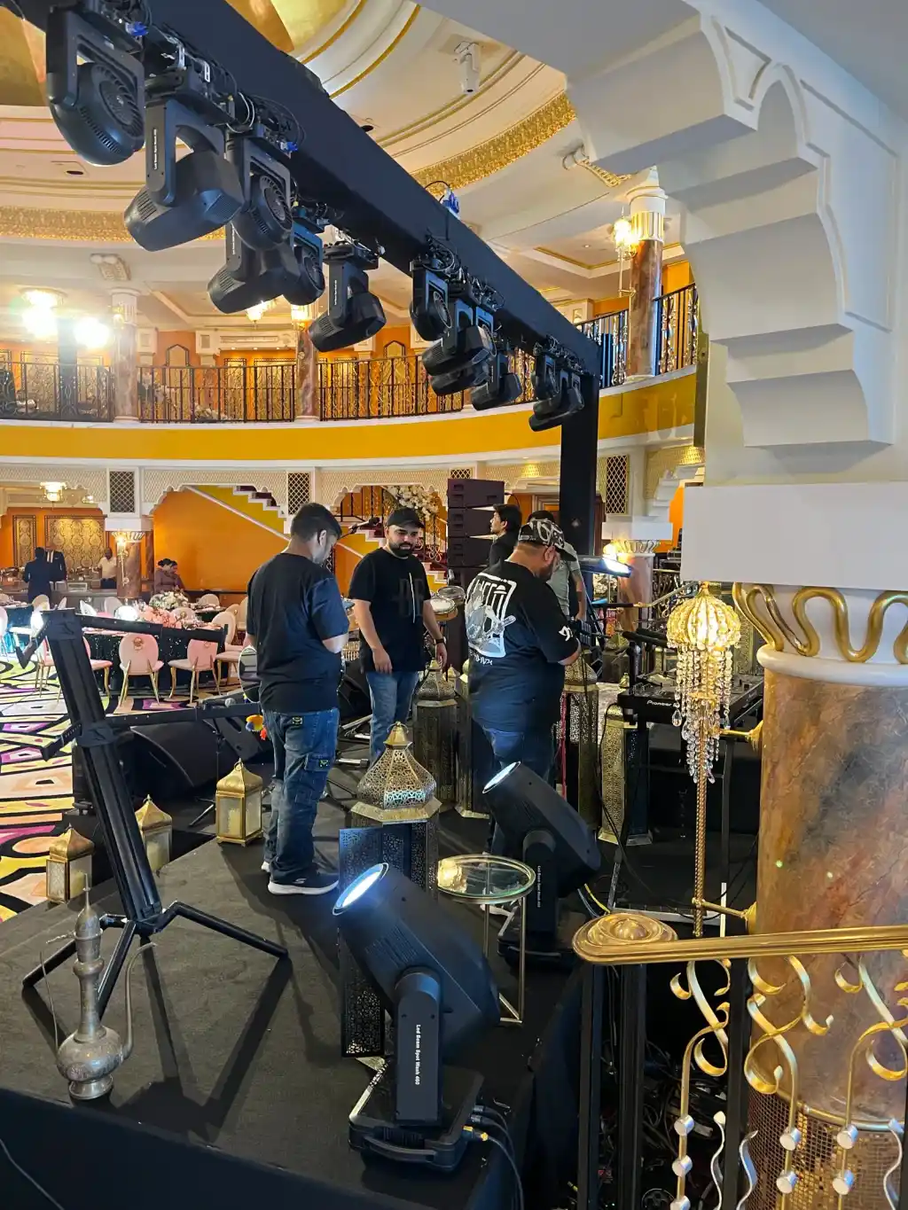 Aayrish Events crew setting brass lanterns across a ballroom stage beneath a truss of moving lights, the gold balcony rising behind them