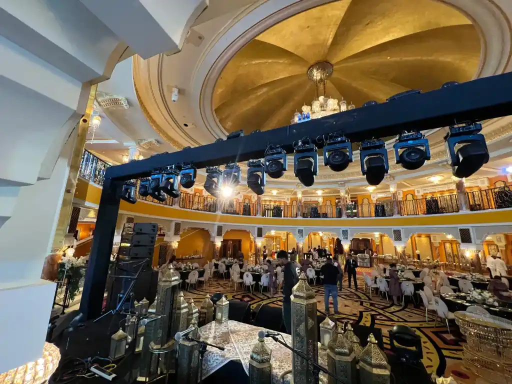 A domed ballroom set for a banquet, with a lighting truss rigged above the stage and a chandelier under a gold leaf dome