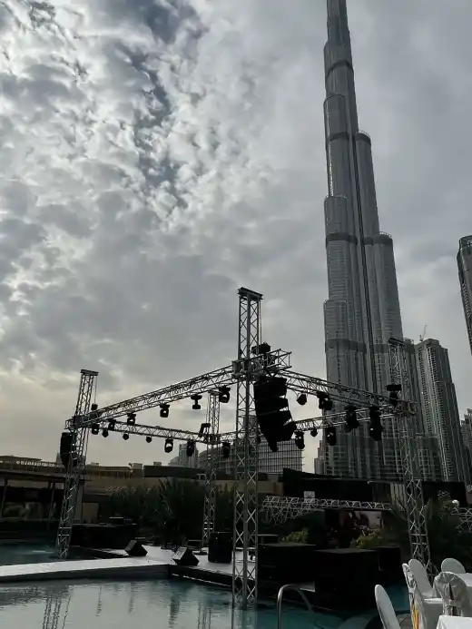 A poolside stage rig of lighting truss and line array speakers standing over a hotel pool, city towers behind under a cloudy sky