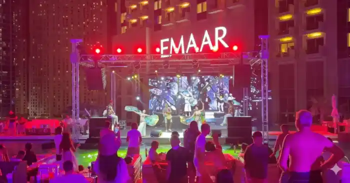 A poolside stage with dancers and an LED screen, lit red and purple, with a crowd watching from the water's edge