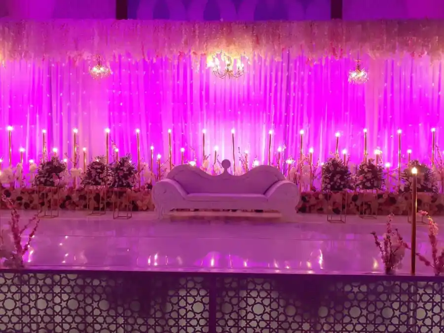A wedding stage backed by sheer curtains lit deep pink, hung with cascading white blossom and small chandeliers, a carved white settee centred between banks of roses and tapered candles