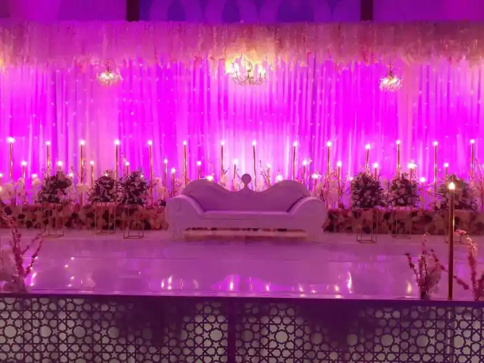 A wedding stage backed by sheer curtains lit deep pink, hung with cascading white blossom and small chandeliers, a carved white settee centred between banks of roses and tapered candles